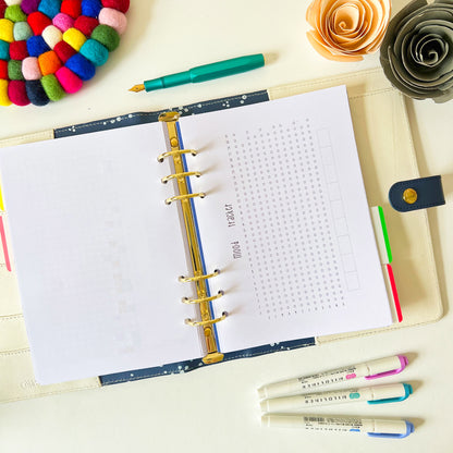 An open Yearly Tracker Planner Page with metal rings lies on a table, featuring a grid and word search puzzle. Surrounding it are colorful pens, highlighters, and decorative items, including a multicolored pom-pom coaster, paper flowers, and a mood a day tracker.