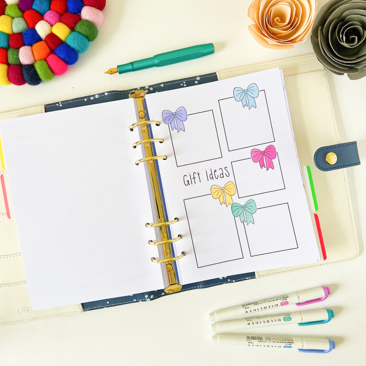 An open planner lies on a white surface, displaying a "Gift Ideas" page with five boxes, each adorned with a colorful bow. Nearby are pens, a green fountain pen, a Birthday Calendar for seamless birthday planning, and two decorative paper flowers, one orange and one grey.