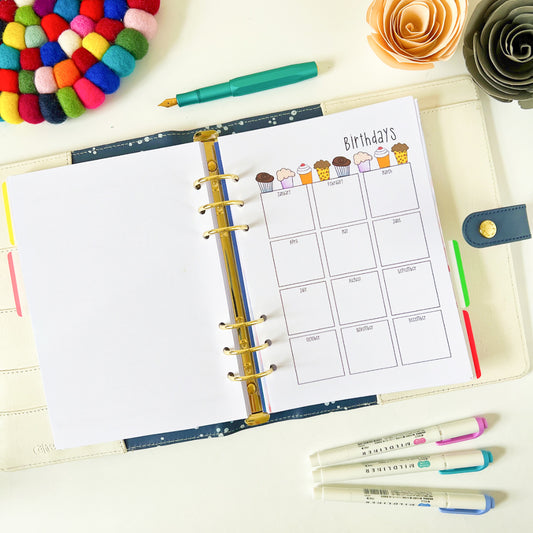 A neatly organized Birthday Calendar is open on a desk, displaying a "Birthdays" page adorned with cupcake illustrations for each month. Surrounding the Birthday Calendar are colorful pens, a multicolored pom-pom coaster, and two paper flowers in yellow and grey—a perfect setup for birthday planning and gift ideas.
