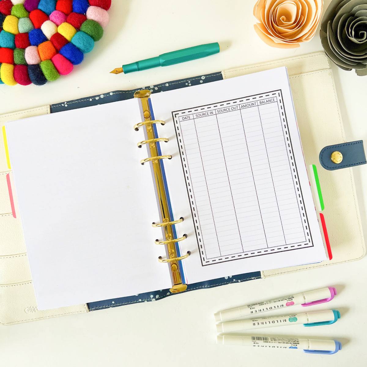 An open planner with a blank Spending Tracker Page inside sits on a white surface. Nearby are colorful pens, a pen, a gray paper flower, and a colorful pom-pom coaster. The planner is surrounded by bright and cheerful stationery items, perfect for detailed budget tracking.