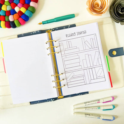 An open Reading Log lies on a table with a colorful wool pom-pom coaster, paper flowers, and pastel highlighters around it. The planner pages include an illustration of a bookshelf for tracking reading progress. A green pen is placed across the top of the open pages.