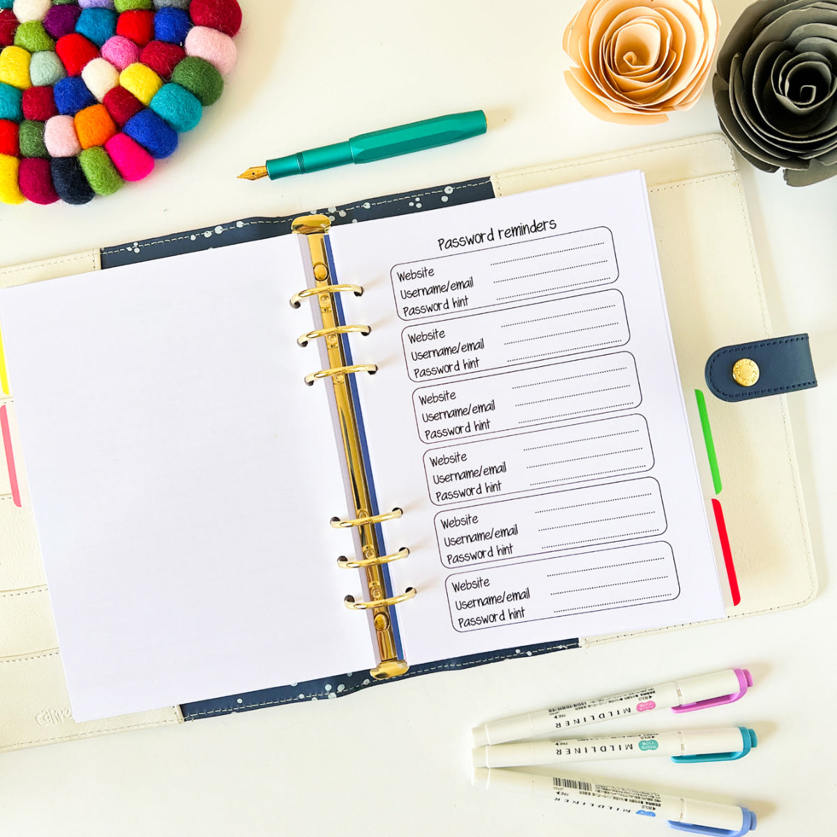An open planner lies on a desk, featuring a Password Keeper page titled “Password reminders” with lines for website, username/email, and password hints. Around it are colorful pens, markers, a paper flower, and a felt ball coaster.