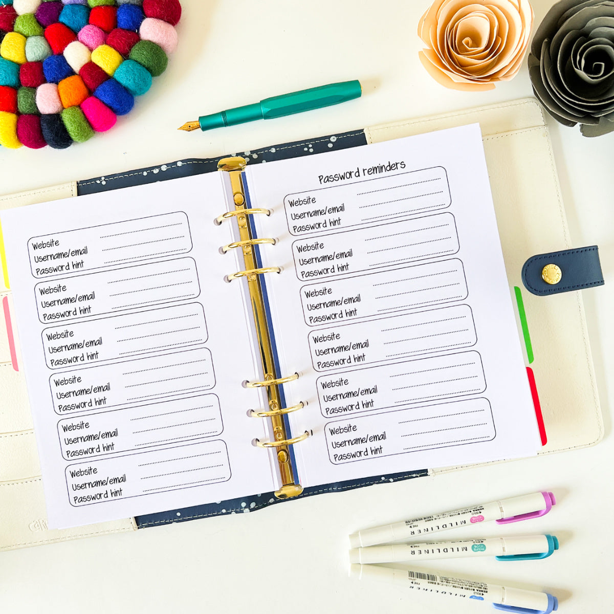 An open planner with a "Password Keeper" page doubles as the perfect password keeper, with sections for website, username/email, and password hints. Surrounding it are colorful accessories: a felt ball coaster, a gold paper flower, a pen, and highlighters.