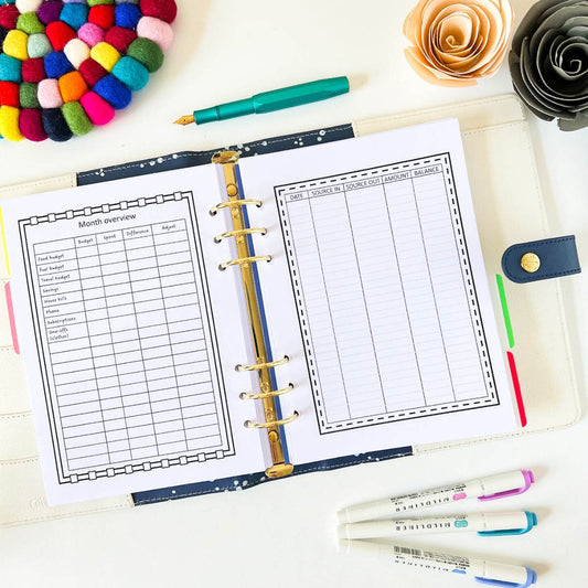 An open Monthly Budget And Spending lies on a desk, displaying pages for monthly overview, daily notes, and budget planning. Nearby are pastel-colored highlighters, a green pen, a multicolored felt ball mat, and decorative paper flowers in orange and cream.