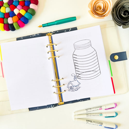 An open planner lies on a white background with a doodle of a jar and a small house with smoke coming out of its chimney on the right-hand page, labeled as a House Deposit Saving Tracker. Nearby are colorful markers, a multicolored felt ball coaster, and two ornamental paper flowers.