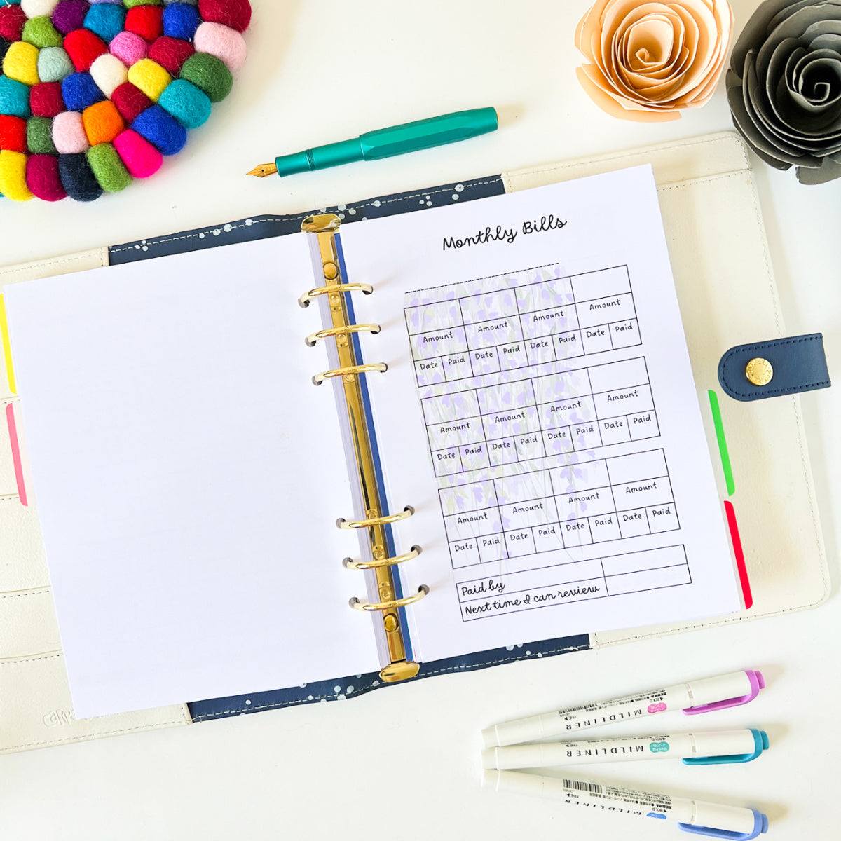 Open planner with "Monthly Bills" sheet on a desk. This Household bill tracker features a six-ring binder holding pages for dates, amounts, and descriptions—perfect for bill tracking. Surrounding it are multicolored pom-pom coaster, pink and yellow flowers, green pen, and pastel-colored highlighters.