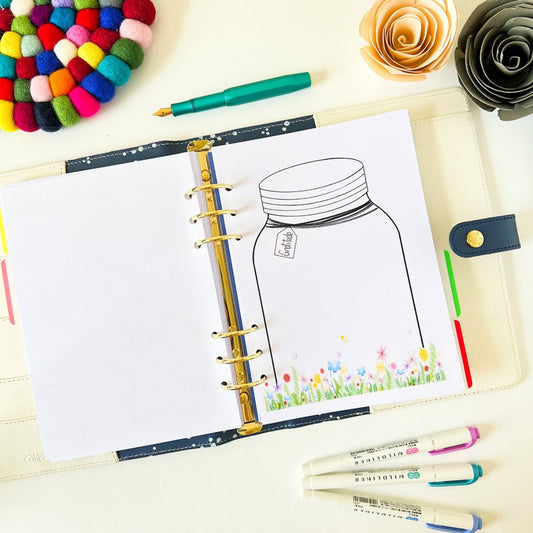 An open "Gratitude Jar" lies on the table, its blank page featuring a gratitude jar illustration amid a vibrant floral border. Nearby are colored markers, a green pen, a multicolored pom-pom coaster, and paper flowers.