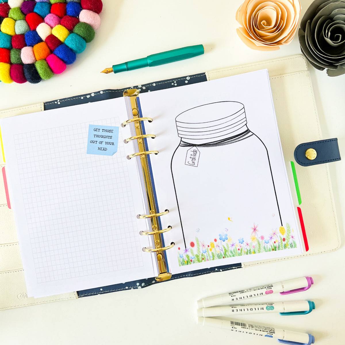 An open notebook with a drawing of a Gratitude Jar and flowers on the right page and a grid pattern with a sticky note saying, "Get those thoughts out of your head" on the left page. Around it are colorful pens, a felt ball coaster, and paper flowers creating a charming floral border.