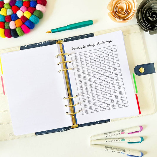 An open Penny Saving Challenge lies on a desk, displaying "Penny Saving Challenge" planner pages. Surrounding the Penny Saving Challenge are colorful pens, highlighters, a felt ball coaster, and paper flowers, creating a decorative and organized workspace.