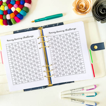 An open Penny Saving Challenge displays two sheets titled "Penny Saving Challenge," each with a 12x5 savings tracker grid containing different monetary amounts. The planner pages are accompanied by colorful pens, markers, a decorative rosette, and a vibrant pom-pom trivet.