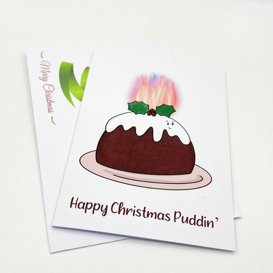 Two greeting cards are displayed. The front card, encased in a festive coloured envelope, is the "Christmas Pudding Christmas Card," featuring an illustration of a flaming Christmas pudding with holly on top and the text "Happy Christmas Puddin'." The second card is partially visible. Both come in a biodegradable cellophane bag to keep them pristine.