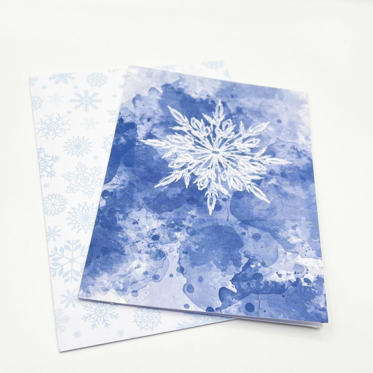 The Snowflake Christmas Card features a blue card adorned with a white snowflake design, placed on top of a white card printed with light blue snowflakes. The festive envelope adds a charming touch, and the card is blank inside for your personal message.