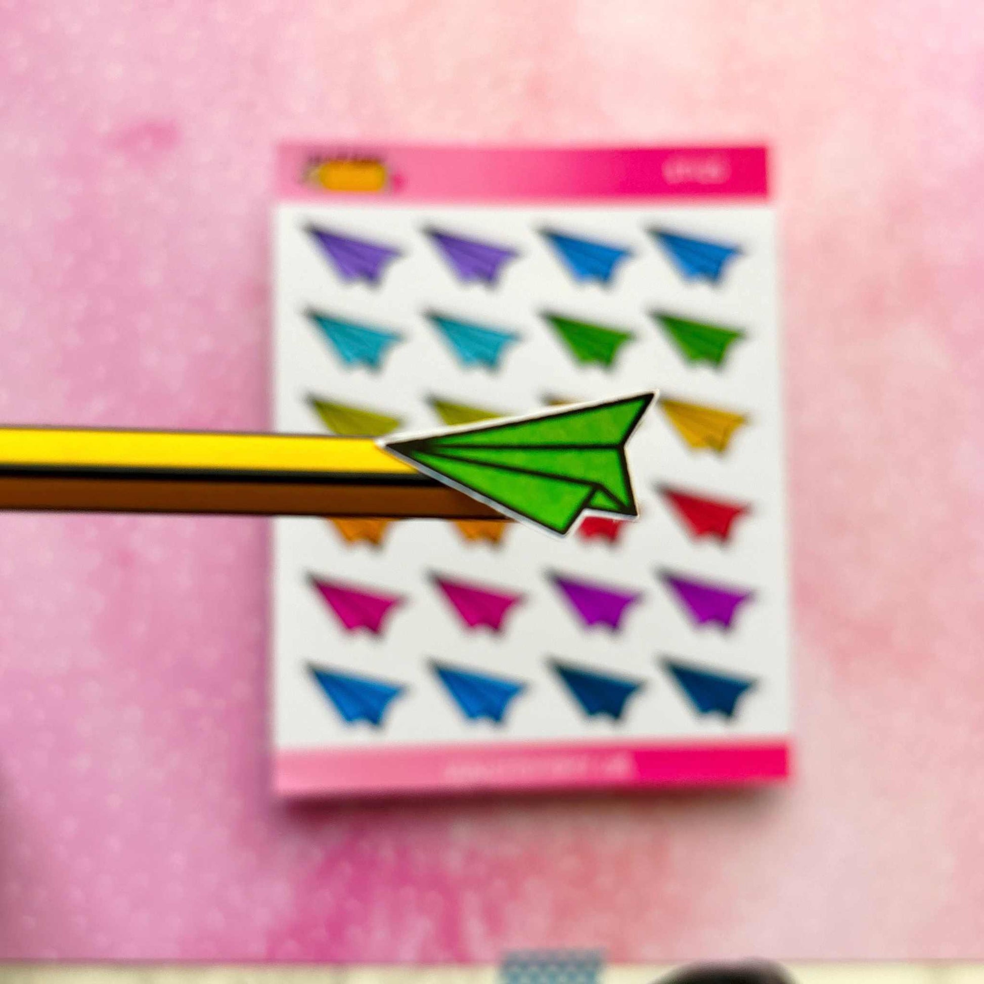 A yellow pencil with the tip wrapped in a green paper airplane shape is showcased alongside the vibrant Paper Aeroplane Sticker Sheet, which features various colorful paper airplane designs, making it perfect for planner decoration.