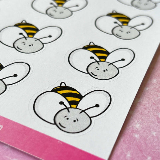 The Bumblebee Sticker Sheet showcases cartoon bumblebee faces with white wings and a yellow stripe pattern, all set against a pink surface.