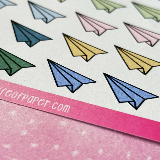 Close-up of the Paper Aeroplane Sticker Sheet, showcasing colorful paper airplanes in blue, yellow, pink, green, and gray on a pink background with partially visible text, evoking a nostalgic sense of fun.