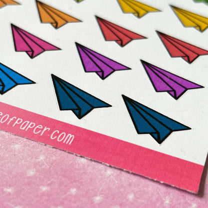 A close-up photo showcasing the Paper Aeroplane Sticker Sheet, adorned with vibrant stickers in blue, purple, orange, and yellow hues on a pink backdrop. Ideal for adding flair to your planner decorations.