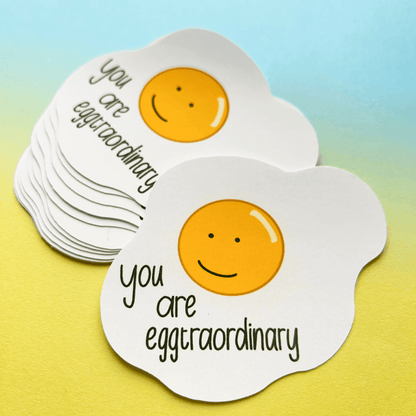 The Extraordinary Silly Pun Quote Vinyl Sticker features a smiling cartoon egg yolk with the phrase "You are eggtraordinary" set against a gradient yellow and blue background. These waterproof vinyl stickers make the perfect gift for friends!