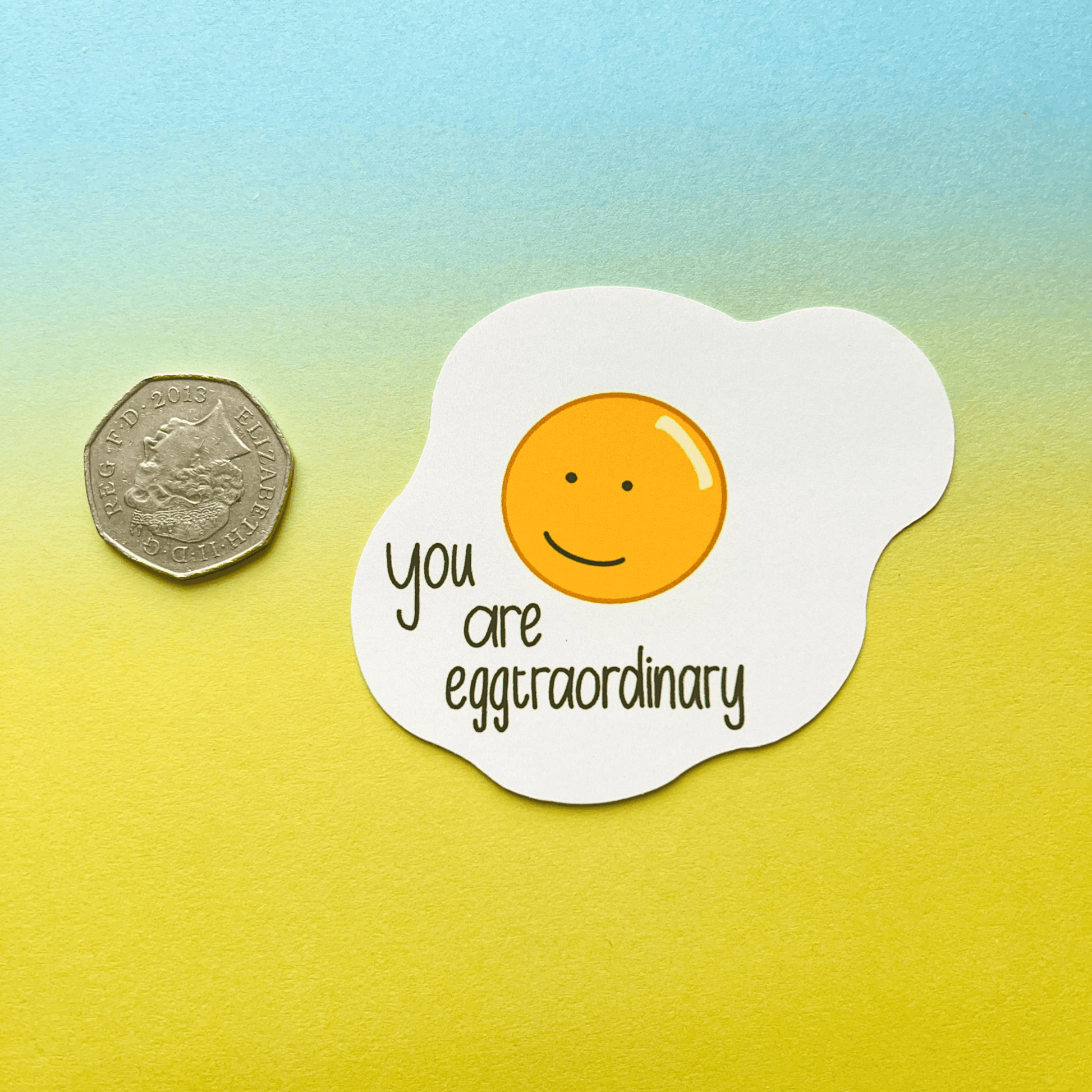 A 50 pence coin and the "Extraordinary Silly Pun Quote Vinyl Sticker," a waterproof vinyl decal shaped like a sunny-side-up egg with a smiling face and the text "you are eggtraordinary" on a yellow to blue gradient background, perfect as a friend gift.