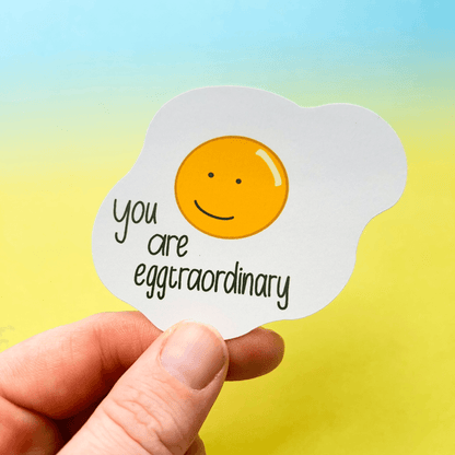 A hand holds the "Extraordinary Silly Pun Quote Vinyl Sticker," which is shaped like a fried egg with a smiling face and features the text "you are eggtraordinary," making it the perfect friend gift.