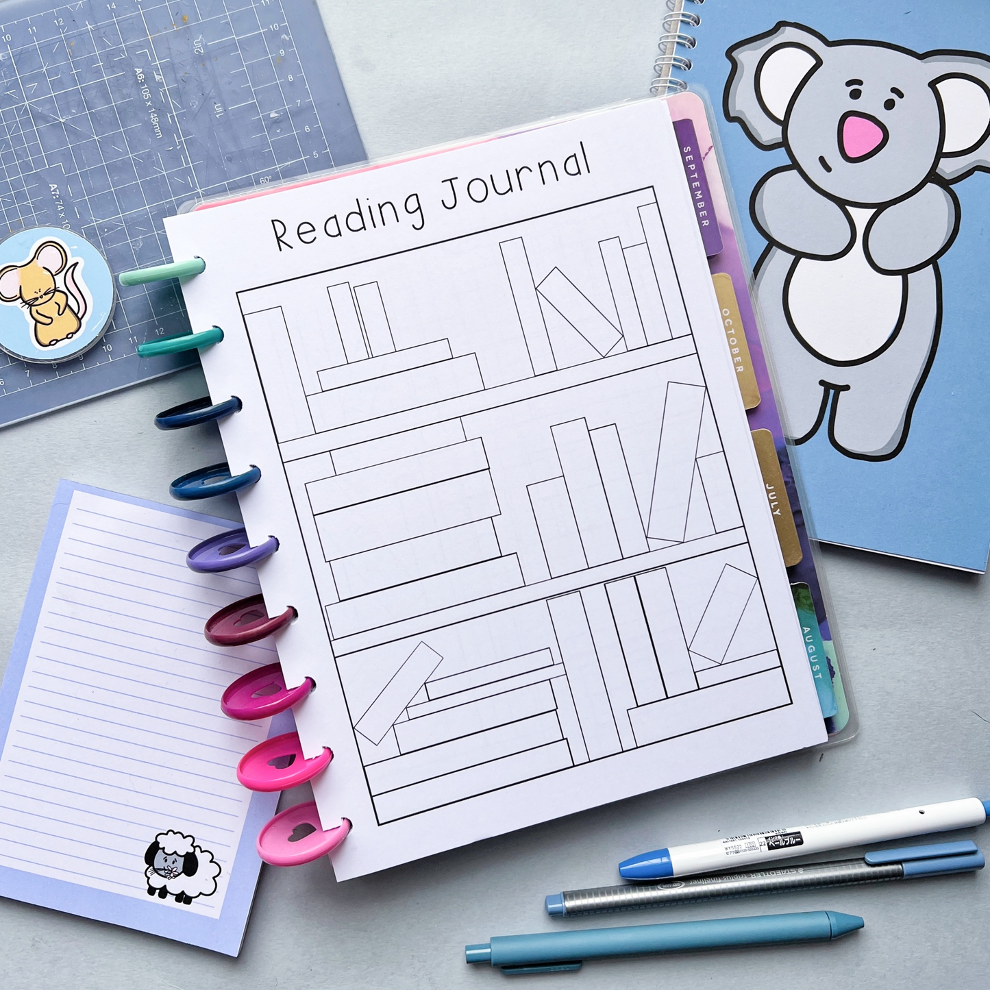 An open binder labeled "Reading Log" is displayed on a white surface. The page shows an outline of bookshelves, perfect for your book tracker. Surrounding the binder are colorful highlighters, a pen, a multicolored pom-pom coaster, and paper flowers.