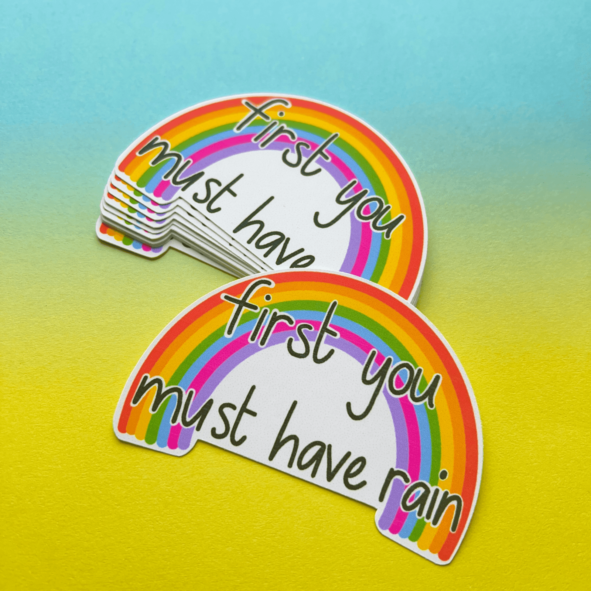 A stack of Rainbow Vinyl Stickers featuring a vibrant rainbow with the text "first you must have rain" on a stunning gradient blue-to-yellow background. These matte, water-resistant vinyl stickers are perfect for adding a splash of color anywhere.