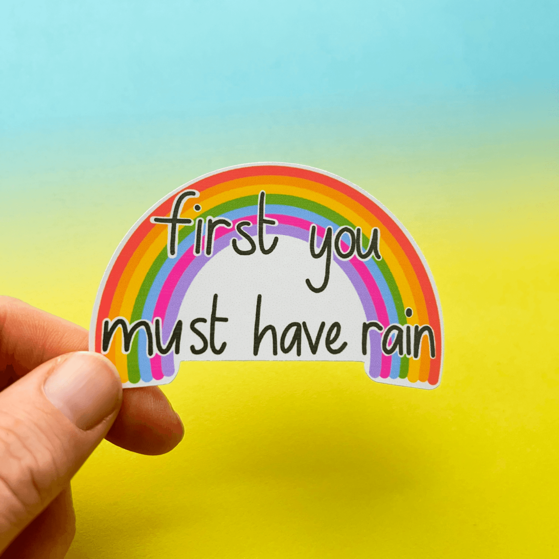 A hand holds a Rainbow Vinyl Sticker, featuring a matte finish with a vibrant rainbow illustration and the text "first you must have rain," against a gradient background of blue and yellow.