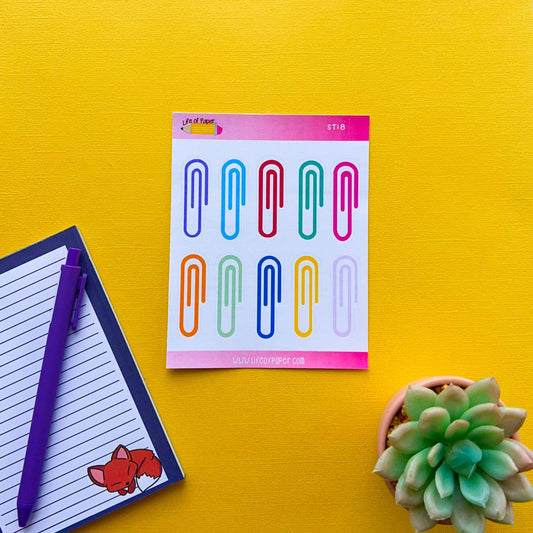 A colorful notepad with Rainbow Paperclip Planner Stickers lies on a bright yellow surface. To the left, there is a small notepad with a purple pen and a fox illustration in the corner, ideal for life planning and task tracking. A green succulent plant is positioned on the right.