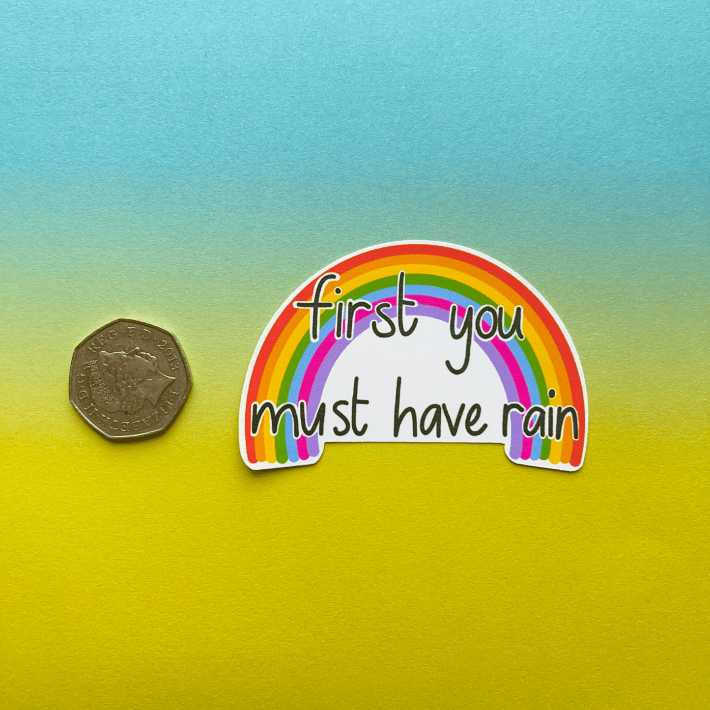 A water-resistant Rainbow Vinyl Sticker with a vibrant rainbow and the text "first you must have rain" displayed next to a coin for size reference, set against a gradient blue to yellow background. This charming sticker is made of high-quality matte vinyl, ensuring durability and bright colors.