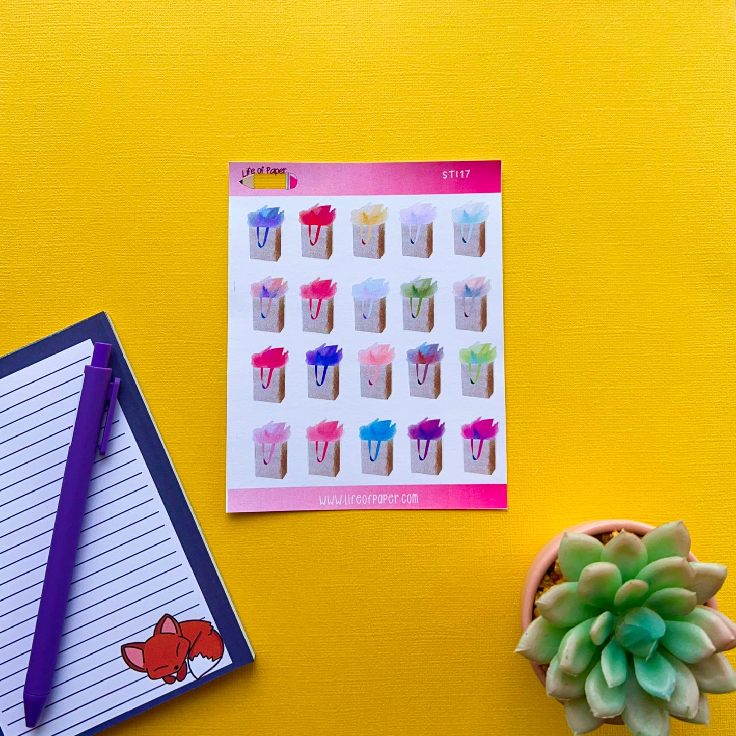 A bright yellow background showcases a sheet of Gift Bag Planner Stickers depicting various colorful drinks with straws. To the left, there's a lined notebook with a fox sticker and two purple pens. A succulent plant is situated in the bottom right corner.
