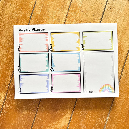 An A4 desk weekly planner pad lies on a wooden surface featuring colorful boxes for each day, ideal for organizing your weekly tasks. A vibrant rainbow at the bottom-right corner adds a cheerful touch to your organized plan.