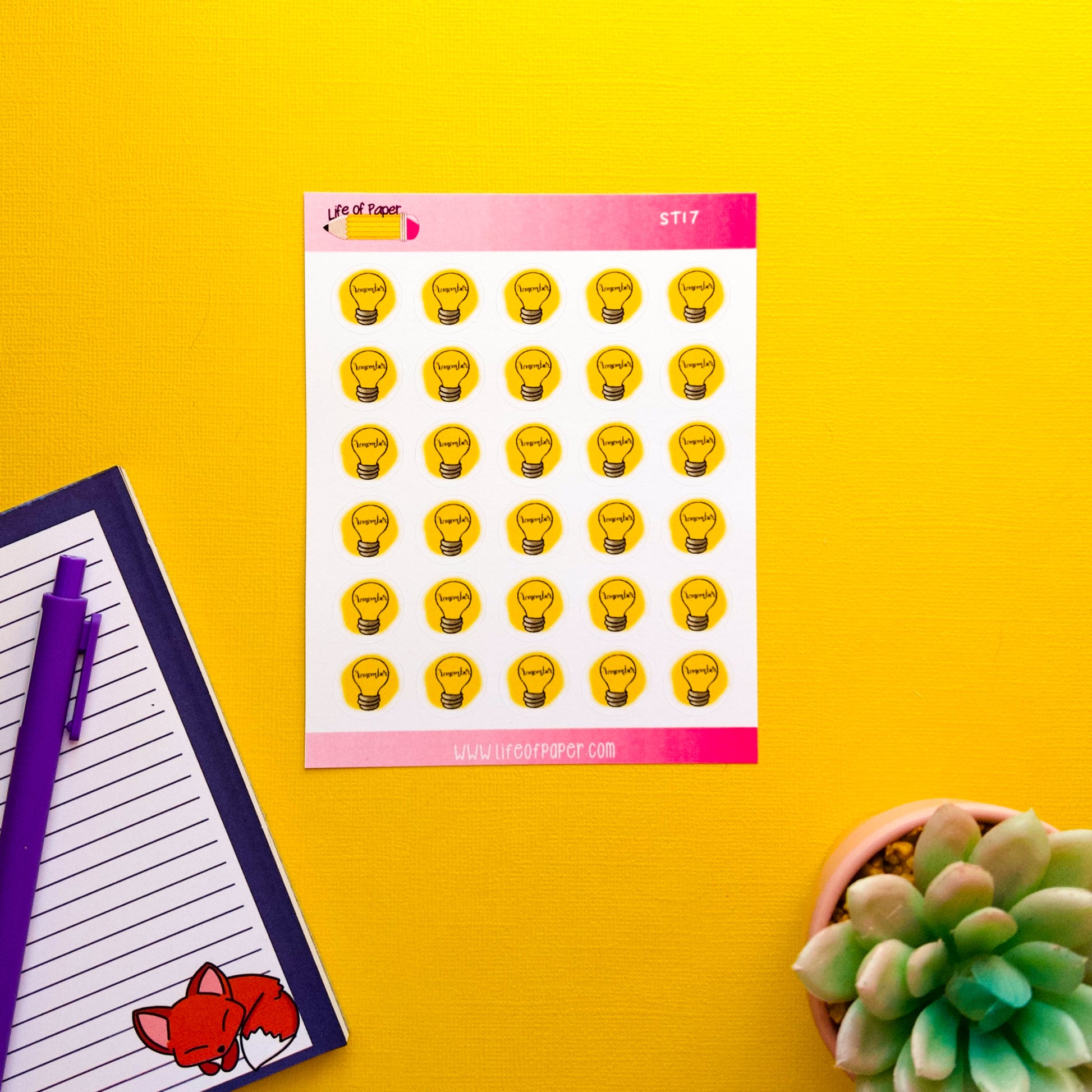 A sheet of Remember Planner Stickers featuring various expressions against a bright yellow background is on the left, along with a notepad with a fox illustration and a purple pen, perfect for planning your goals. A small potted succulent plant is on the right.
