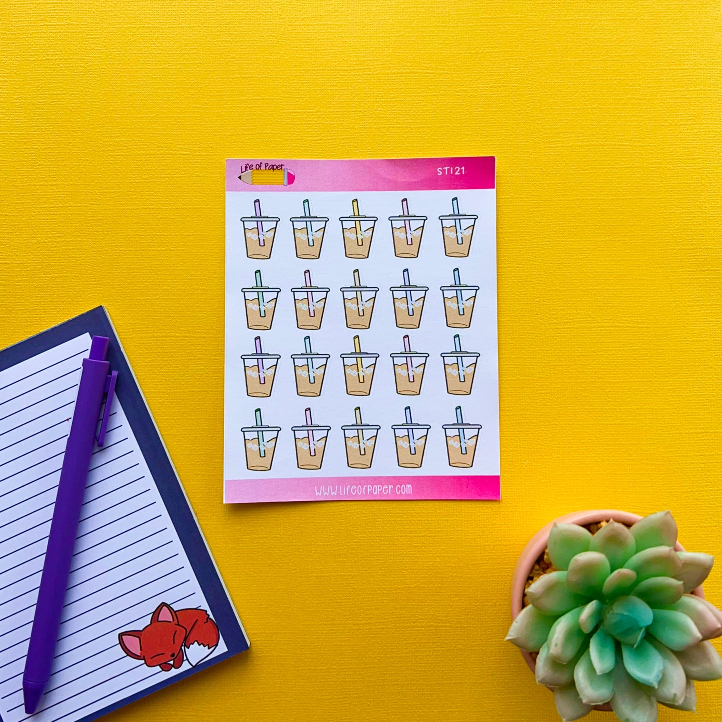 A notepad with Iced Coffee Planner Stickers is on a bright yellow surface. Next to the notepad is a lined notebook with a purple pen resting on it and a small drawing of a fox in the corner. A green succulent plant is partially visible on the right.