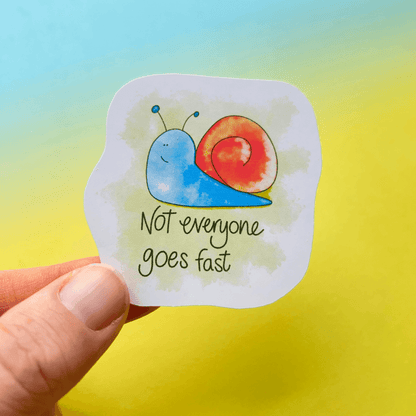 A hand holds the vibrant Speedy Snail - Animal Vinyl Sticker, showcasing a water-resistant vinyl design with a charming snail illustration and the text, "Not everyone goes fast," set against a gradient background transitioning from yellow to blue. This colourful decal adds both charm and durability to any surface.