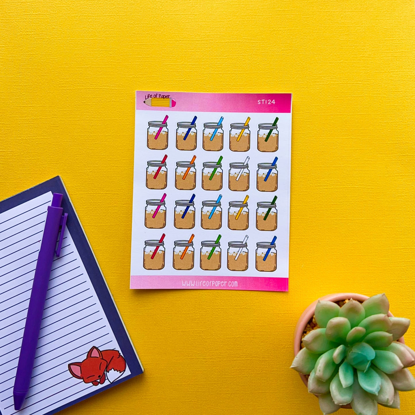 Colorful sheet of Mason Coffee Jar Planner Stickers with vibrant designs attached to a yellow board. To the left, there is a striped notepad adorned with a charming fox illustration and two pens placed on top. At the bottom right, there is a small, green succulent plant adding a touch of nature.