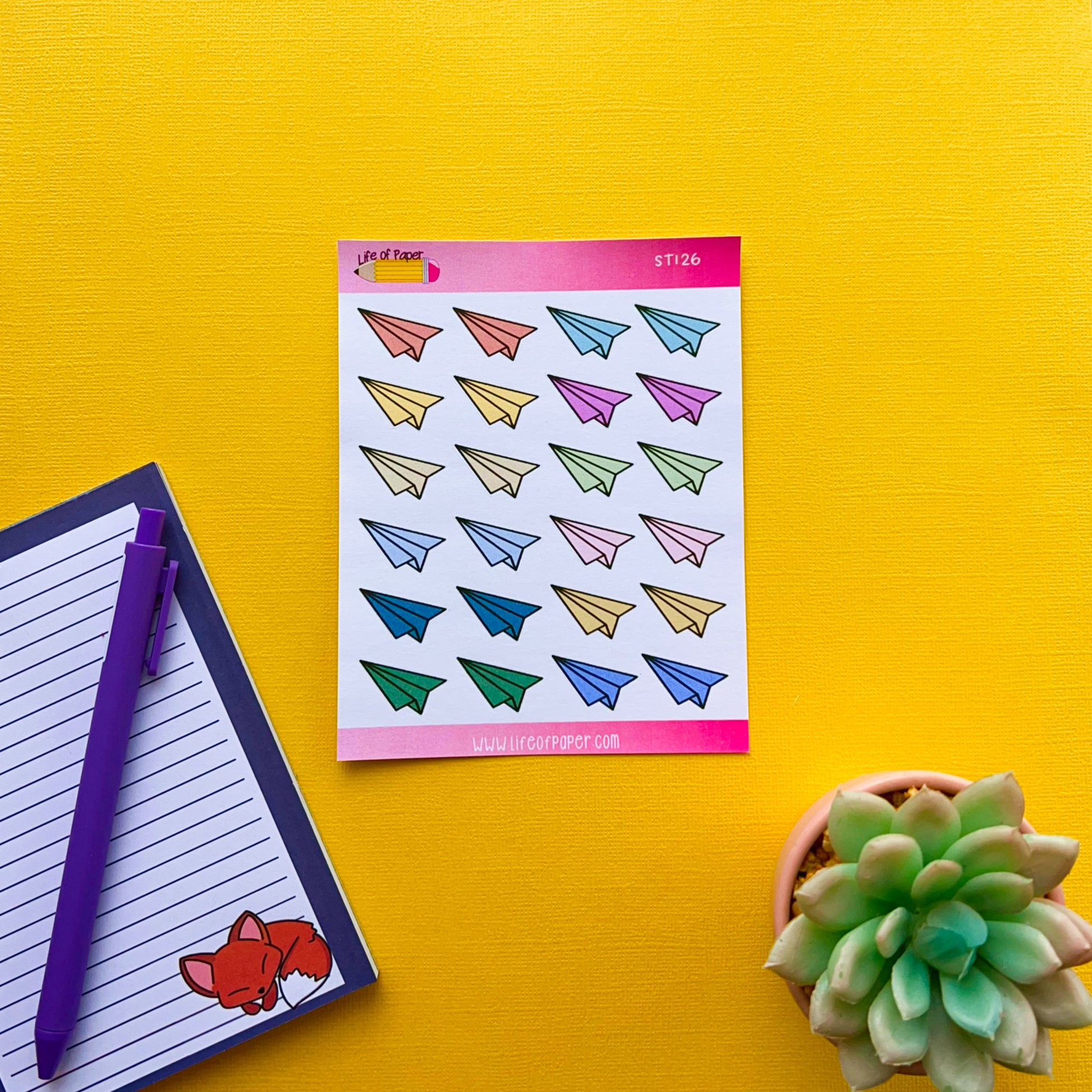 A colorful Paper Aeroplane Sticker Sheet lies on a bright yellow surface. To the left, there is a notebook with a purple pen resting on it, featuring a small fox sticker in the corner. On the right, a green succulent plant is placed in a small pot, adding to the nostalgic fun.
