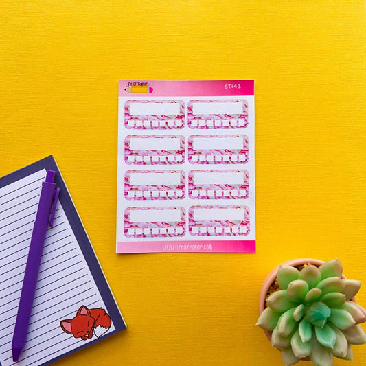 A sheet of rectangular Weekly Habit Tracker Planner Stickers with pink borders on a yellow surface. Next to it is a notepad with a sleeping fox illustration in the corner, accompanied by a purple pen. A small green succulent is placed on the right side.