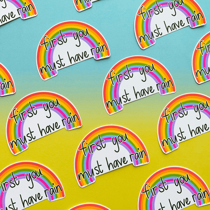 Introducing the Rainbow Vinyl Sticker, displayed on a gradient background and featuring rainbow designs with the text "first you must have rain." These water-resistant stickers boast durability and charm, making them perfect for any surface.