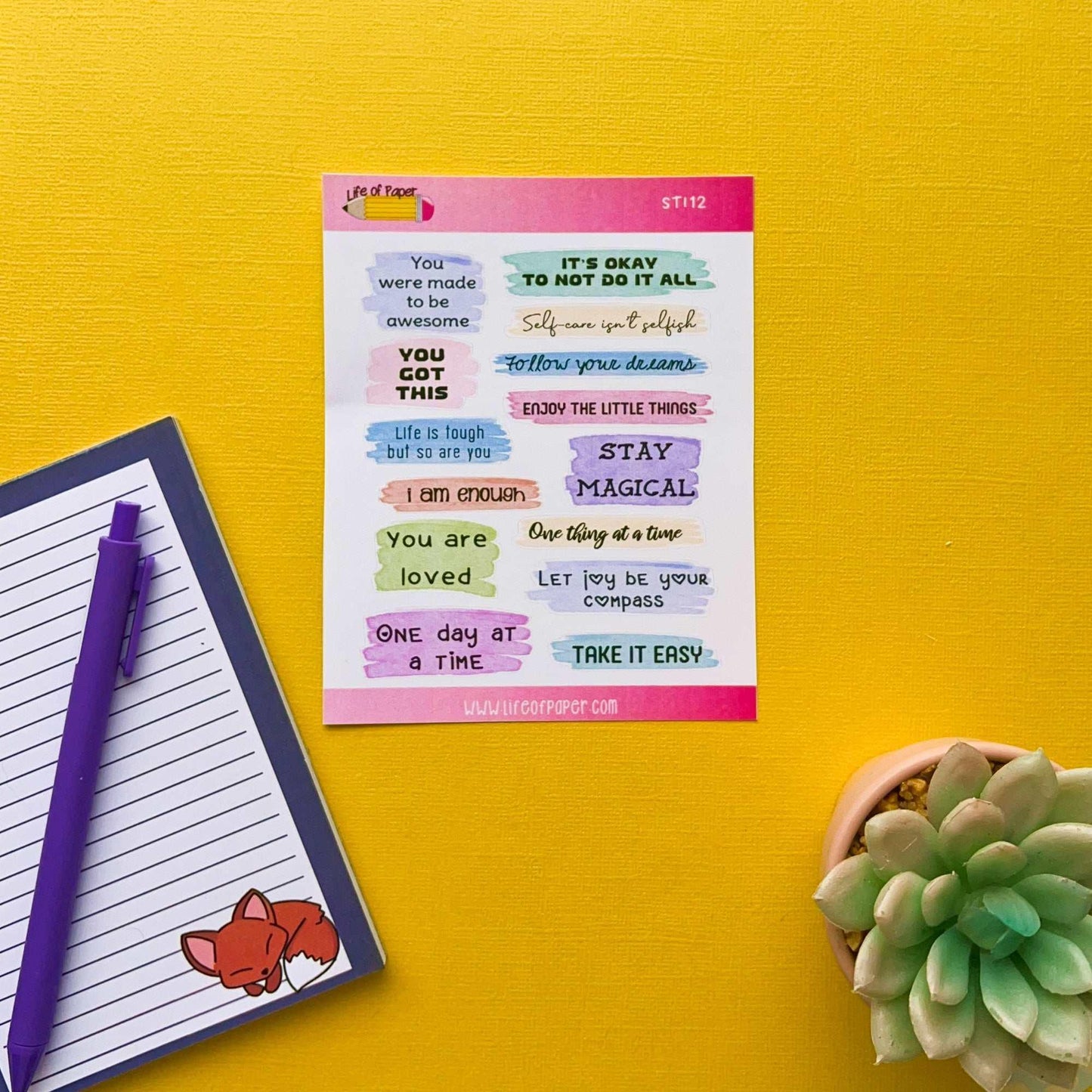 A colorful sheet of Positive Quote Planner Stickers on a yellow background, featuring phrases like "You got this," "Stay magical," and "Take it easy." Nearby, an open notepad with a fox illustration and a purple pen are placed alongside a small succulent plant.