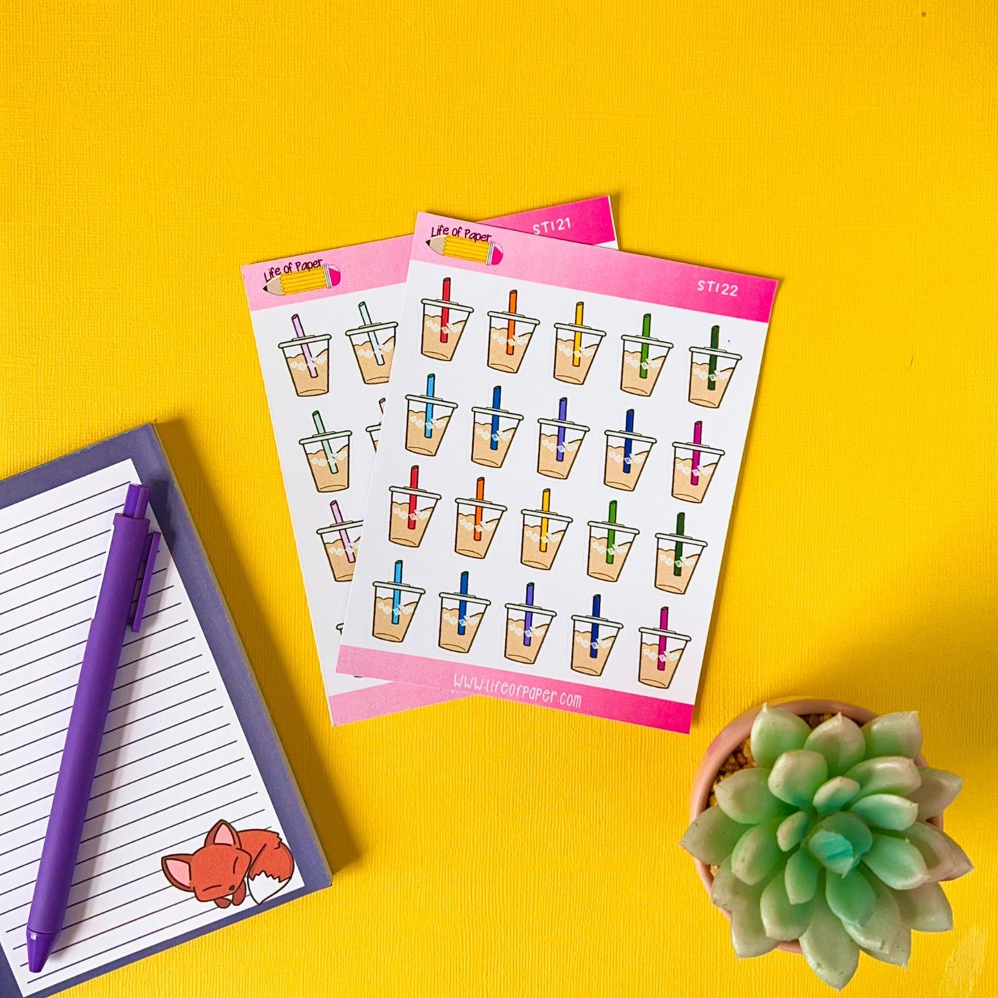 Two sheets of Iced Coffee Stickers, a notepad with a fox illustration and a purple pen, and a small potted succulent are placed on a bright yellow surface.