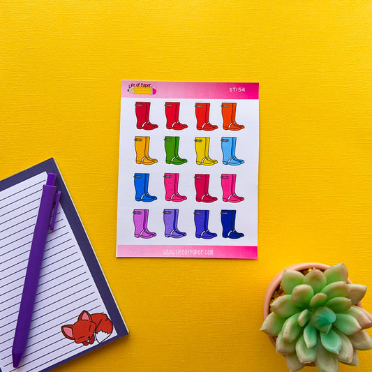 A vibrant yellow background showcases a sheet of colorful Wellington Boot Planner Stickers arranged in four rows with different colors. Below the stickers are a notepad with a fox illustration, a purple pen, and a small potted succulent plant.
