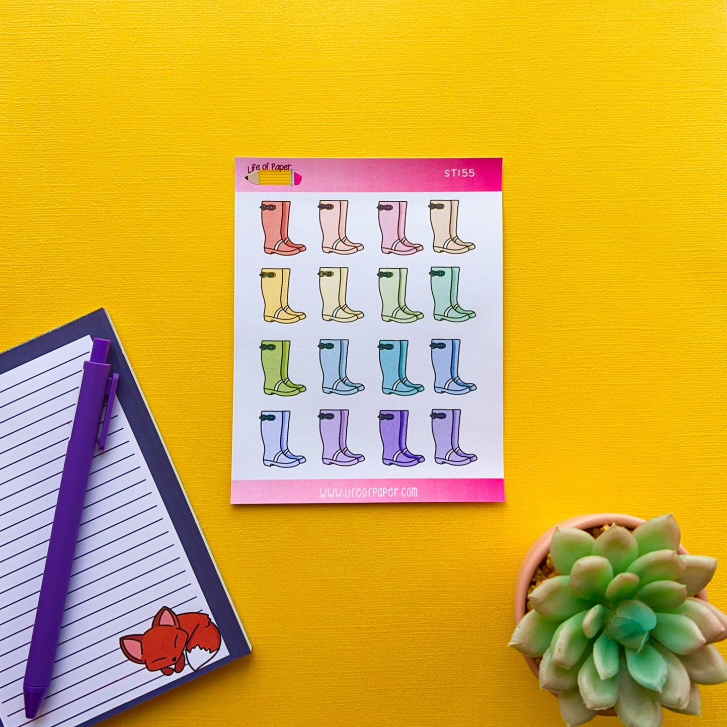 A yellow surface holds a notepad showing colorful Wellington Boot Planner Stickers in rows, with each pair in a different color. To the left is a pen next to a lined notepad with a fox illustration. A small succulent plant is at the bottom right corner.