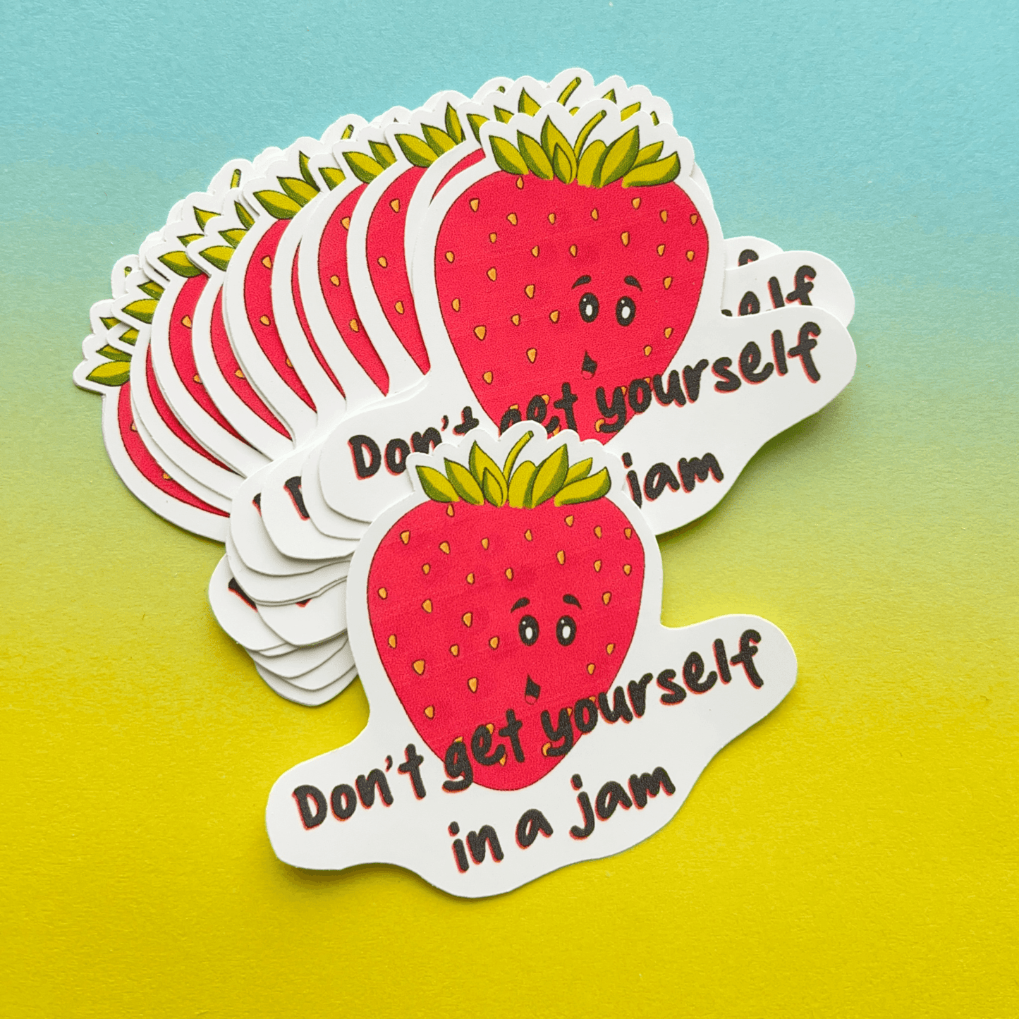 The Strawberry Sticker is a water-resistant vinyl decal showcasing a cheerful cartoon strawberry with a smiling face and the playful pun "Don't get yourself in a jam" set against a gradient background.