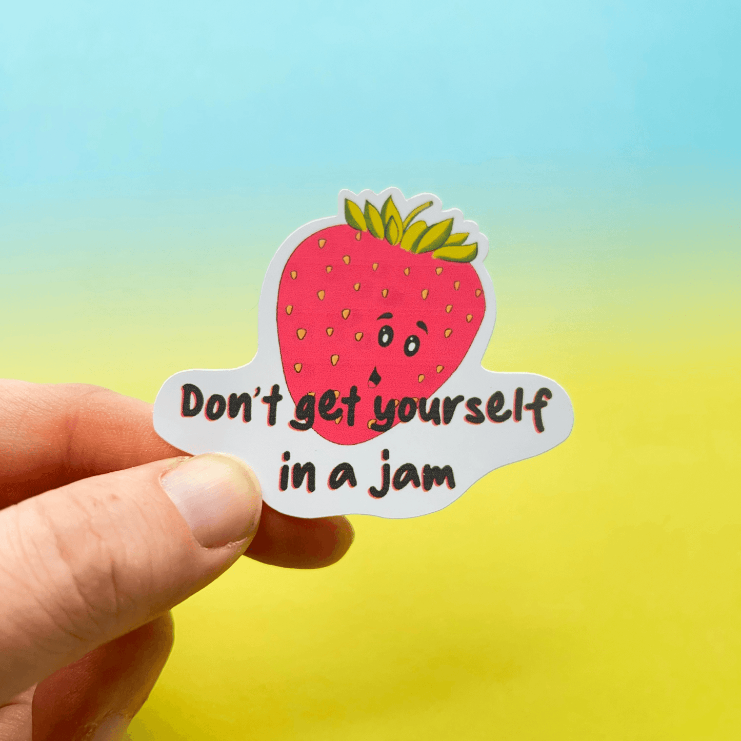 A hand holds the Strawberry Sticker, a vinyl depiction of a cartoon strawberry with a face and the text "Don't get yourself in a jammy pun," set against a gradient blue and yellow background. This water-resistant gem is perfect for adding some playful charm wherever you stick it.