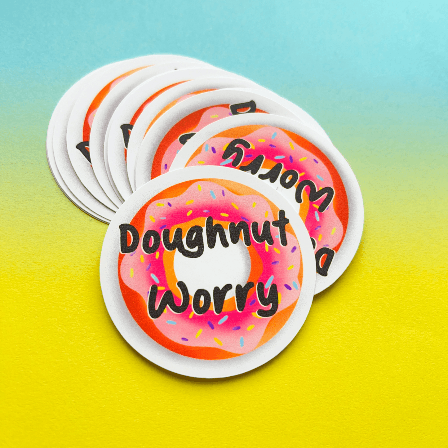 The Doughnut Sticker showcases a pink doughnut image coupled with the playful phrase "Doughnut Worry" on a gradient blue and yellow backdrop. Crafted from durable matte vinyl, these water-resistant stickers are an ideal way to add a whimsical touch to your possessions.