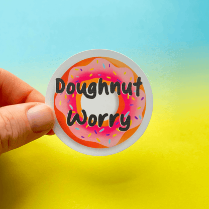 Hand holding the Doughnut Sticker, featuring a pink frosted doughnut graphic with the text "Doughnut Worry". The circular sticker, crafted from strong matte vinyl and water-resistant, is set against a gradient background of blue to yellow.