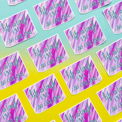 The "Life Is Tough Affirmation Vinyl Sticker" is showcased in a grid pattern on a gradient surface. This supportive decal, featuring the phrase "Life is tough but so are you" on a purple and pink abstract background, is made from strong matte vinyl and includes positive quotes.