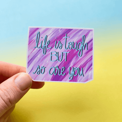 A hand holds a small, durable matte Life Is Tough Affirmation Vinyl Sticker with a purple and pink background that reads, "life is tough BUT so are you.