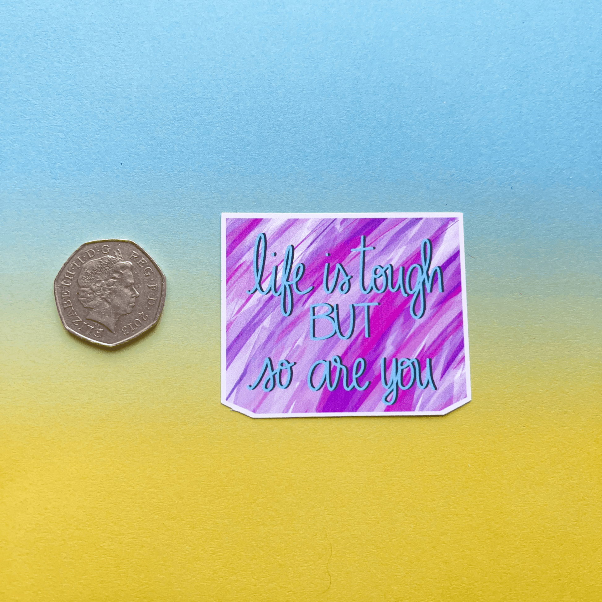 A coin next to a Life Is Tough Affirmation Vinyl Sticker with the message "Life is tough but so are you" on a blue and yellow gradient background, mimicking the look of a supportive decal.