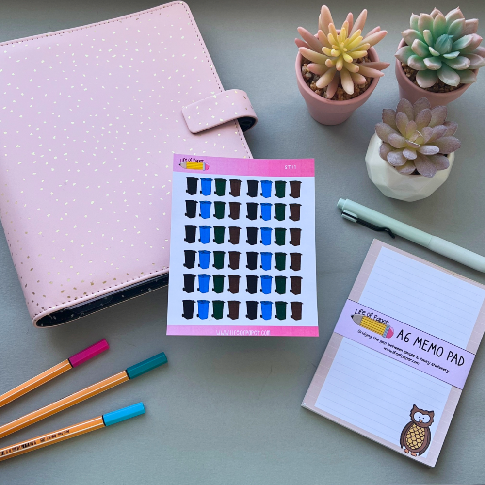 A pastel pink planner sits on a desk beside colorful pens, a white pen, a memo pad featuring an owl, and Wheelie Bin Planner Stickers with blue and black rain boots. Various small succulent plants in pots are arranged in the background.