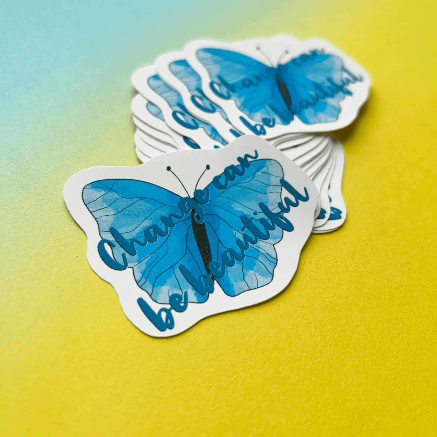 The "Change can be Beautiful Vinyl Sticker" showcases a blue butterfly illustration along with the inspirational text "Change can be beautiful," all set against a gradient yellow to blue background. These matte vinyl stickers are perfect for adding a touch of positivity to your space.